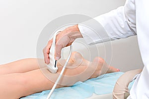 Doctor in white uniform with diagnostic equipment performs an ultrasound using a device on the legs of a female patient. Surgeon