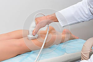 Doctor in white uniform with diagnostic equipment performs an ultrasound using a device on the legs of a female patient. Surgeon