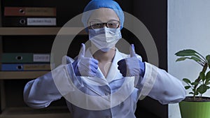 Doctor In White Uniform Shows Thumbs Up Sign. Female Medical Worker In Protective Mask And Disposable Gloves Shows