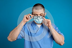 Doctor wiping foggy glasses caused by wearing disposable mask on blue background. Protective measure during coronavirus pandemic