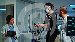 Doctor witing on clipboard while patient with mask running on cross trainer