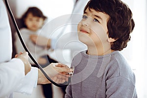 Doctor-woman examining a child patient by stethoscope. Cute arab boy and his brother at physician appointment. Medicine