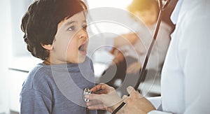 Doctor-woman examining a child patient by stethoscope in sunny clinik. Cute arab boy and his brother at physician