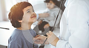 Doctor-woman examining a child patient by stethoscope in sunny clinik. Cute arab boy and his brother at physician