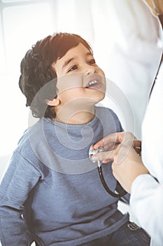 Doctor-woman examining a child patient by stethoscope in sunny clinik. Cute arab boy and his brother at physician
