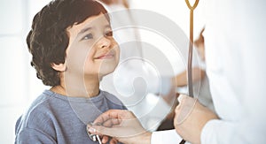 Doctor-woman examining a child patient by stethoscope in sunny clinik. Cute arab boy and his brother at physician