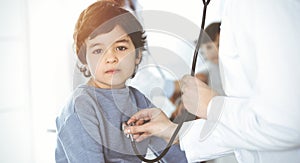 Doctor-woman examining a child patient by stethoscope in sunny clinik. Cute arab boy and his brother at physician