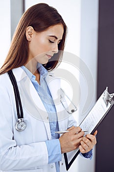 Doctor woman filling up medical form while standing near window in clinic. Physician at work. Medicine and health care