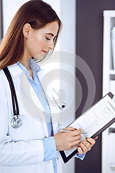 Doctor woman filling up medical form while standing near window in clinic. Physician at work. Medicine and health care