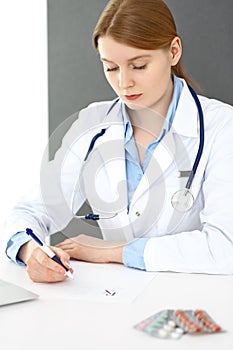 Doctor woman filling up prescription or medical record form while sitting near window in clinic or hospital. Medicin