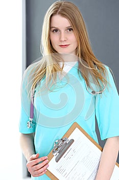 Doctor woman portrait with stethoscope. Young female surgeon or nurse standing near grey wall and window in clinic