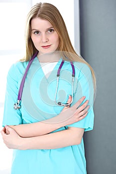 Doctor woman portrait with stethoscope. Young female surgeon or nurse standing near grey wall and window in clinic