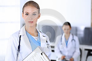 Doctor woman standing in emergency hospital office with colleague at background. Physician at work, studio portrait