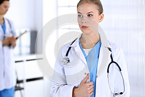 Doctor woman standing in emergency hospital office with colleague at background. Physician at work, studio portrait