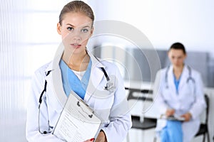 Doctor woman standing in emergency hospital office with colleague at background. Physician at work, studio portrait