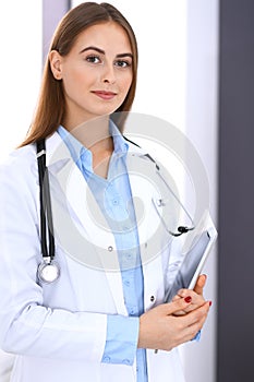 Doctor woman using tablet computer while standing straight near window in hospital. Happy physician at work. Medicine