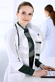 Doctor woman at work with patient and colleague at background. Physician filling up medical documents or prescription