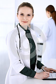 Doctor woman at work with patient and colleague at background. Physician filling up medical documents or prescription