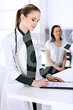 Doctor woman at work with patient and colleague at background. Physician filling up medical documents or prescription