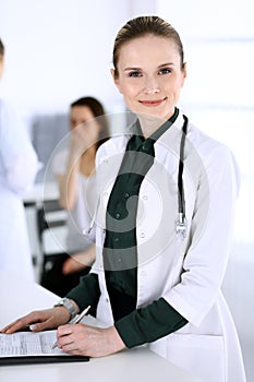 Doctor woman at work with patient and colleague at background. Physician filling up medical documents or prescription