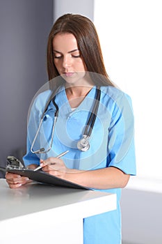 Doctor woman at work. Portrait of female physician filling up medical form while standing near reception desk at clinic