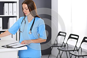 Doctor woman at work. Portrait of female physician filling up medical form while standing near reception desk at clinic