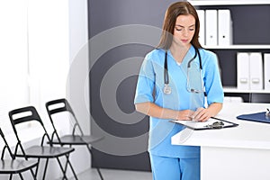 Doctor woman at work. Portrait of female physician filling up medical form while standing near reception desk at clinic