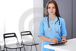 Doctor woman at work. Portrait of female physician filling up medical form while standing near reception desk at clinic