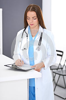 Doctor woman at work. Portrait of female physician filling up medical form while standing near reception desk at clinic