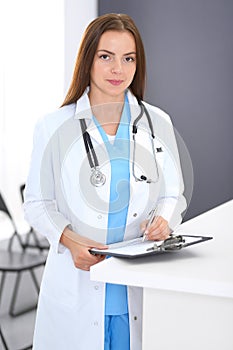 Doctor woman at work. Portrait of female physician filling up medical form while standing near reception desk at clinic