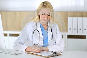 Doctor woman at work while sitting at the desk in hospital or clinic. Blonde cheerful physician filling up medical