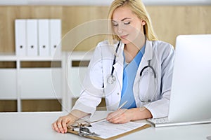 Doctor woman at work while sitting at the desk in hospital or clinic. Blonde cheerful physician filling up medical