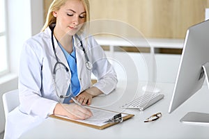 Doctor woman at work while sitting at the desk in hospital or clinic. Blonde cheerful physician filling up medical