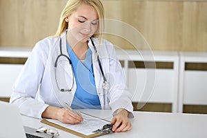 Doctor woman at work while sitting at the desk in hospital or clinic. Blonde cheerful physician filling up medical