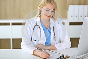 Doctor woman at work while sitting at the desk in hospital or clinic. Blonde cheerful physician filling up medical