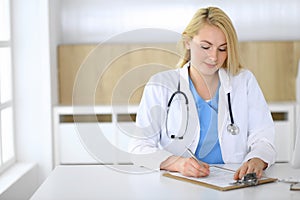 Doctor woman at work while sitting at the desk in hospital or clinic. Blonde cheerful physician filling up medical