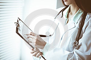 Doctor woman working with medication history records form while standing near window in clinic. Medicine and health care