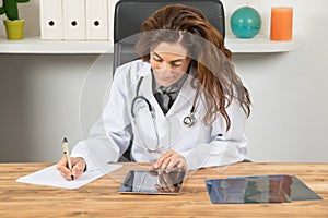 Doctor woman writing on paper looking digital tablet in office