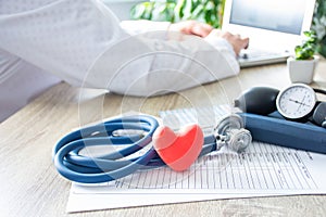 Doctor at work on laptop in blurry background, in foreground - shape of red heart, stethoscope,  sphygmomanometer on medical docum