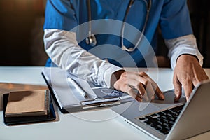 Doctor working on laptop computer and tablet and medical stethoscope on clipboard on desk.