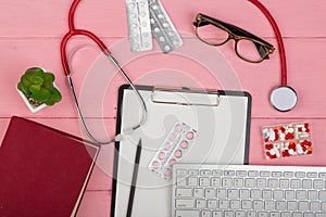 Doctor workplace with blank clipboard, medicine red stethoscope, computer keyboard, eyeglasses, book and pills on pink wooden tabl