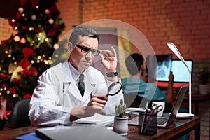 The doctor works on New Year`s Eve. He holds the magnifying glass in his hands. Behind him works a colleague.