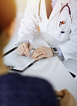 A doctor is writing a medical prescription for his patient, while sitting together at the desk in the sunny cabinet in a