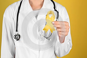 Doctor with yellow ribbon on color background, closeup. Cancer awareness concept