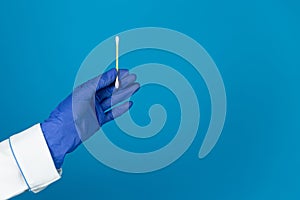 Doctors hand in a glove holds a plastic cotton swab on a blue background with copy space