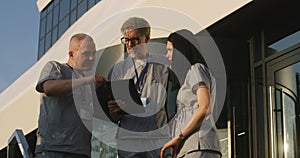 Doctors in uniform stand outdoors at the hospital entrance, use digital tablet