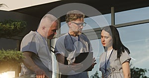 Doctors in uniform stand outdoors at the hospital entrance, use digital tablet