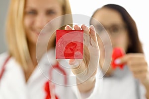 Doctors in white uniforms with red ribbon on chest hold condom in hand closeup