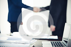 Documents and laptop on the table. Business people shaking hands on the background, silhouettes