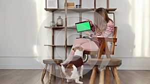 A dog in a brown sweater and a girl look at a screen with chromakey. A woman shows a tablet to a Jack Russell